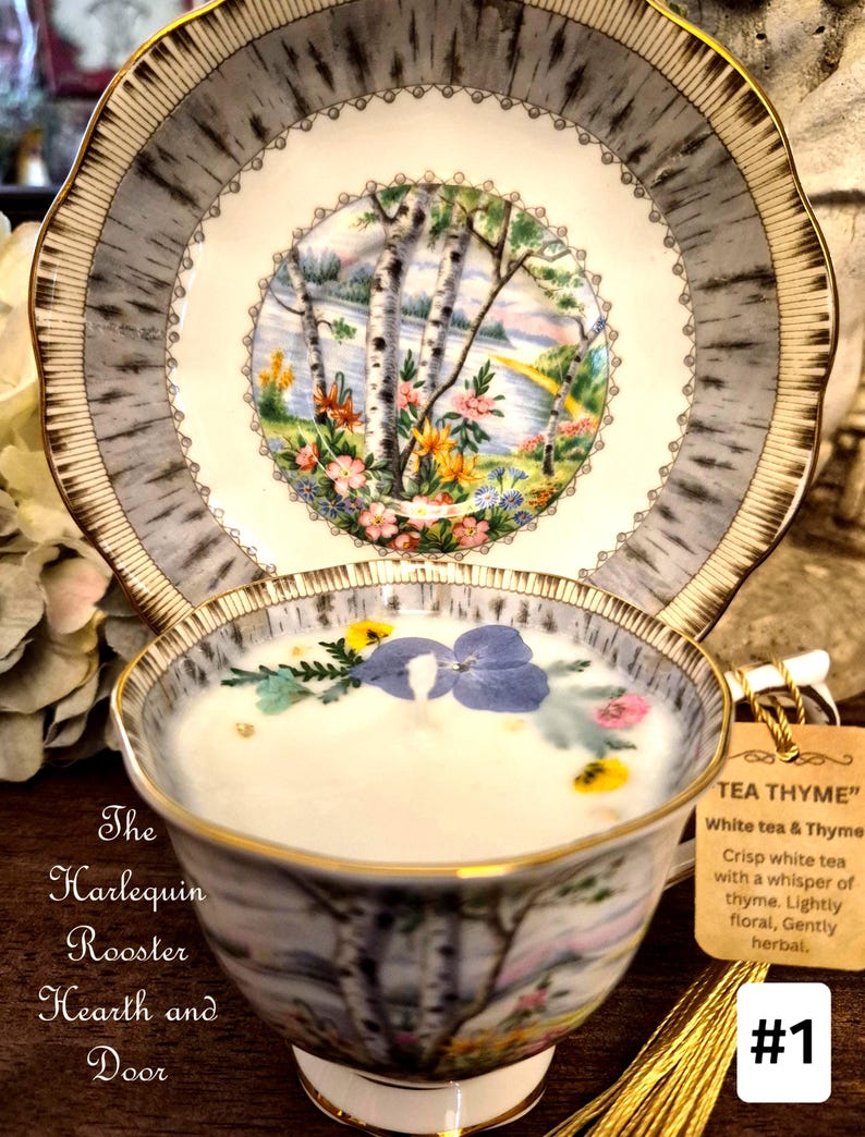May include: A teacup and saucer set with a birch tree design, filled with a white liquid and decorated with flowers. The saucer and cup have gold trim. A tag reads "TEA THYME" with a description of the tea. The text "The Harlequin Rooster Hearth and Door" is also visible.