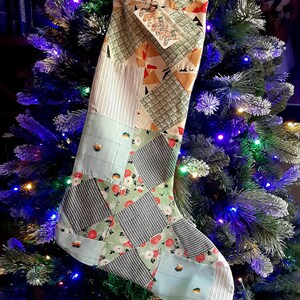 May include: A handmade Christmas stocking with a patchwork design. The stocking features various fabric patterns, including geometric shapes, floral prints, and stripes, in shades of green, red, and blue. It is decorated with small embellishments and hangs on a Christmas tree.