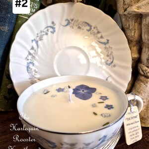 May include: A white teacup candle with blue floral accents and a matching saucer. The candle has a white wax with dried flowers and a wick. A tag reads "TEA THYME". The Harlequin Rooster Hearth and Door.