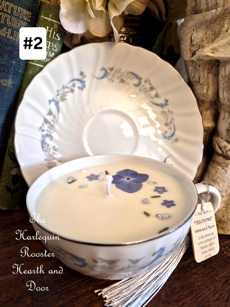 May include: A white teacup candle with blue floral accents and a matching saucer. The candle has a white wax with dried flowers and a wick. A tag reads "TEA THYME". The Harlequin Rooster Hearth and Door.