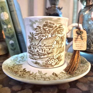 Puede incluir: Un juego de taza de té y platillo de cerámica blanca con un estampado marrón de una casa de campo. Una borla marrón con un dije de llave de metal cuelga del asa, unida a una etiqueta que dice "TEA TIME" y describe té blanco con tomillo.