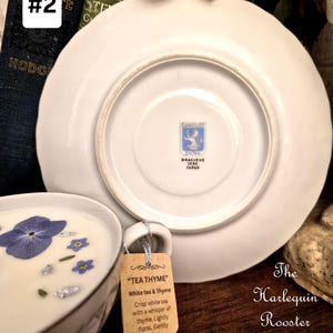 May include: A white teacup and saucer set with a floral design, featuring a tag that reads "TEA THYME". The teacup contains a white liquid with blue flowers and silver flakes. The saucer is white with a blue logo on the bottom. The text "The Harlequin Rooster Hearth and Door" is also visible.