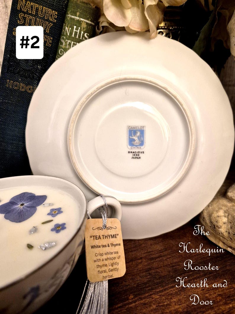May include: A white teacup and saucer set with a floral design, featuring a tag that reads "TEA THYME". The teacup contains a white liquid with blue flowers and silver flakes. The saucer is white with a blue logo on the bottom. The text "The Harlequin Rooster Hearth and Door" is also visible.