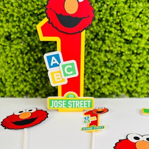 May include: Red Elmo cake topper with a number one, ABC blocks, and a Sesame Street sign that says "JOSE STREET 123".