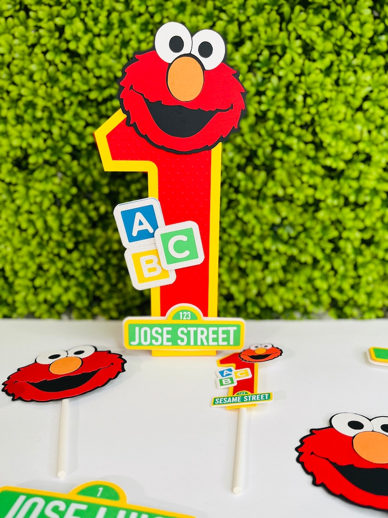 May include: Red Elmo cake topper with a number one, ABC blocks, and a Sesame Street sign that says "JOSE STREET 123".