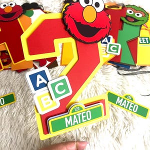 May include: Red number two cake topper with a Sesame Street theme. The number two is decorated with a red Elmo face and yellow and green ABC blocks. The topper also includes a green Sesame Street sign with the name Mateo and the number 123.