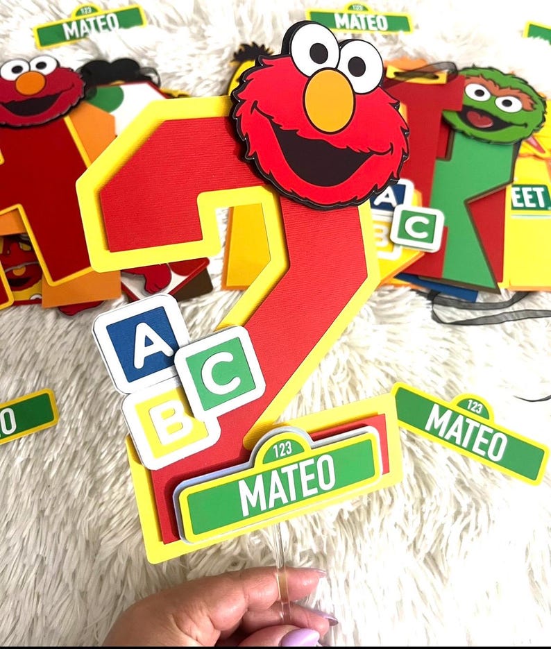 May include: Red number two cake topper with a Sesame Street theme. The number two is decorated with a red Elmo face and yellow and green ABC blocks. The topper also includes a green Sesame Street sign with the name Mateo and the number 123.