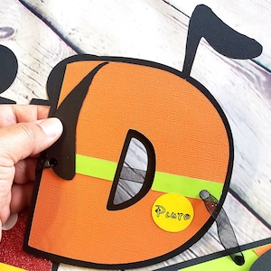 May include: Orange felt letter D with black outline and a green and black stripe. The letter has a black felt ear and a yellow circle with the word "Pluto" written in black.