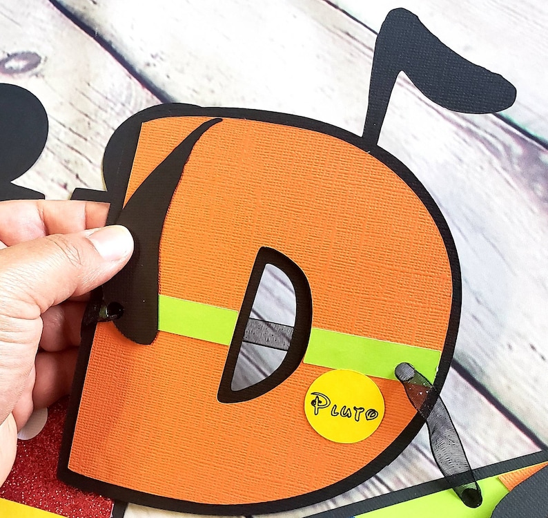 May include: Orange felt letter D with black outline and a green and black stripe. The letter has a black felt ear and a yellow circle with the word "Pluto" written in black.