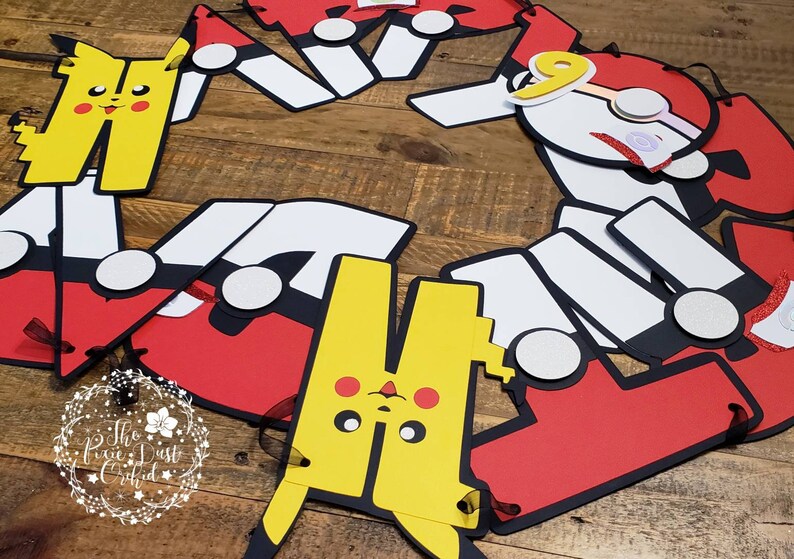 May include: A red, white, and black paper banner with a yellow Pikachu character and a red and white character with a number 9 on it. The banner is decorated with glitter and has black ribbon ties.