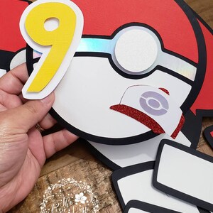 May include: A red and white Pokeball with a black outline and a yellow number 9. The Pokeball has a white center with a silver iridescent circle and a red glitter cap with a white Pokeball symbol.