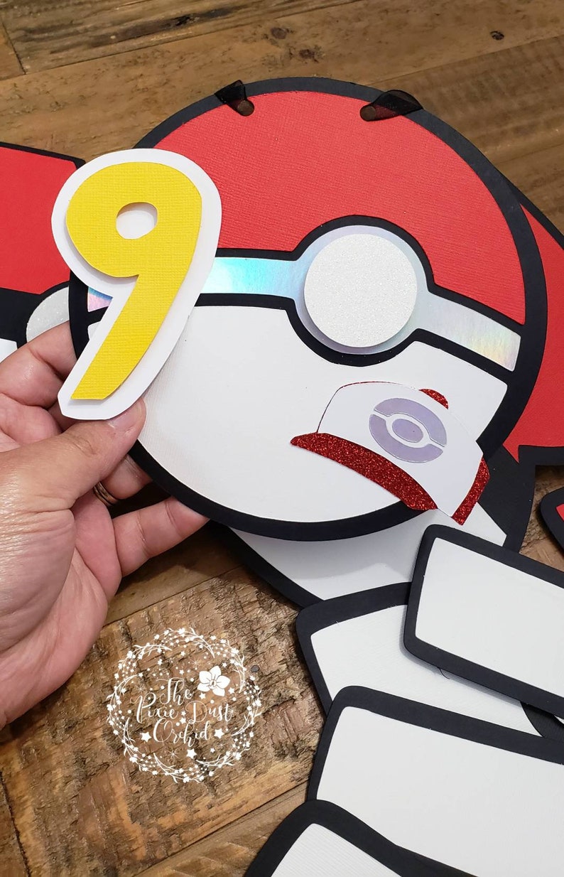 May include: A red and white Pokeball with a black outline and a yellow number 9. The Pokeball has a white center with a silver iridescent circle and a red glitter cap with a white Pokeball symbol.