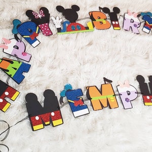 May include: A colorful felt banner with Mickey Mouse characters spelling out "Happy Birthday" and a name. The letters are decorated with glitter and different colors.