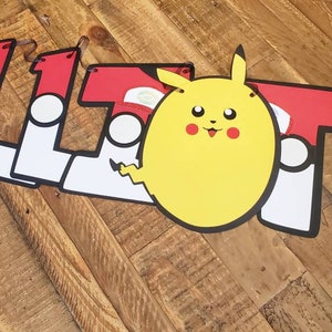 May include: A red, white, and black banner with the name "ELLIOT" spelled out in large letters. Each letter has a white circle with a black outline and a red Pok&eacute; Ball graphic. A yellow Pikachu character is in the center of the banner.