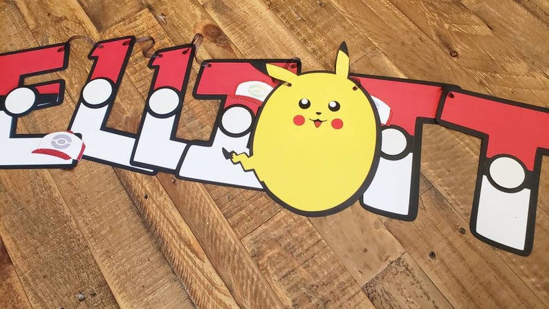 May include: A red, white, and black banner with the name "ELLIOT" spelled out in large letters. Each letter has a white circle with a black outline and a red Pok&eacute; Ball graphic. A yellow Pikachu character is in the center of the banner.