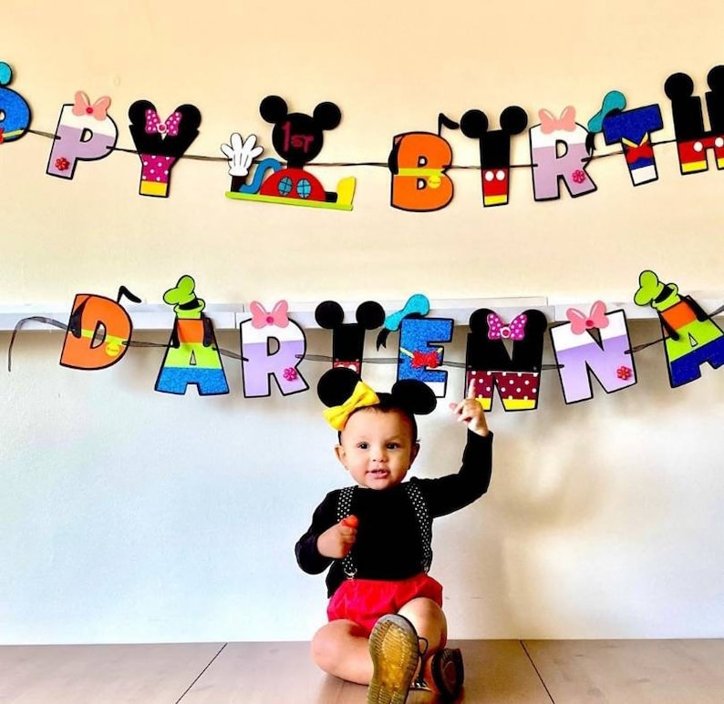 May include: A child wearing a black shirt and red shorts sits in front of a colorful banner that reads "Happy 1st Birthday Darien". The banner features Mickey Mouse and other Disney characters.