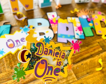 May include: A yellow and purple cake topper with the text "Daniela is One" and a star with colorful sprinkles. The topper is decorated with paint splatters and a yellow duck.