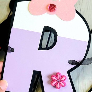 May include: A purple and white letter R with a black outline. The letter has a pink bow with a rhinestone in the center and a pink flower with a rhinestone in the center.