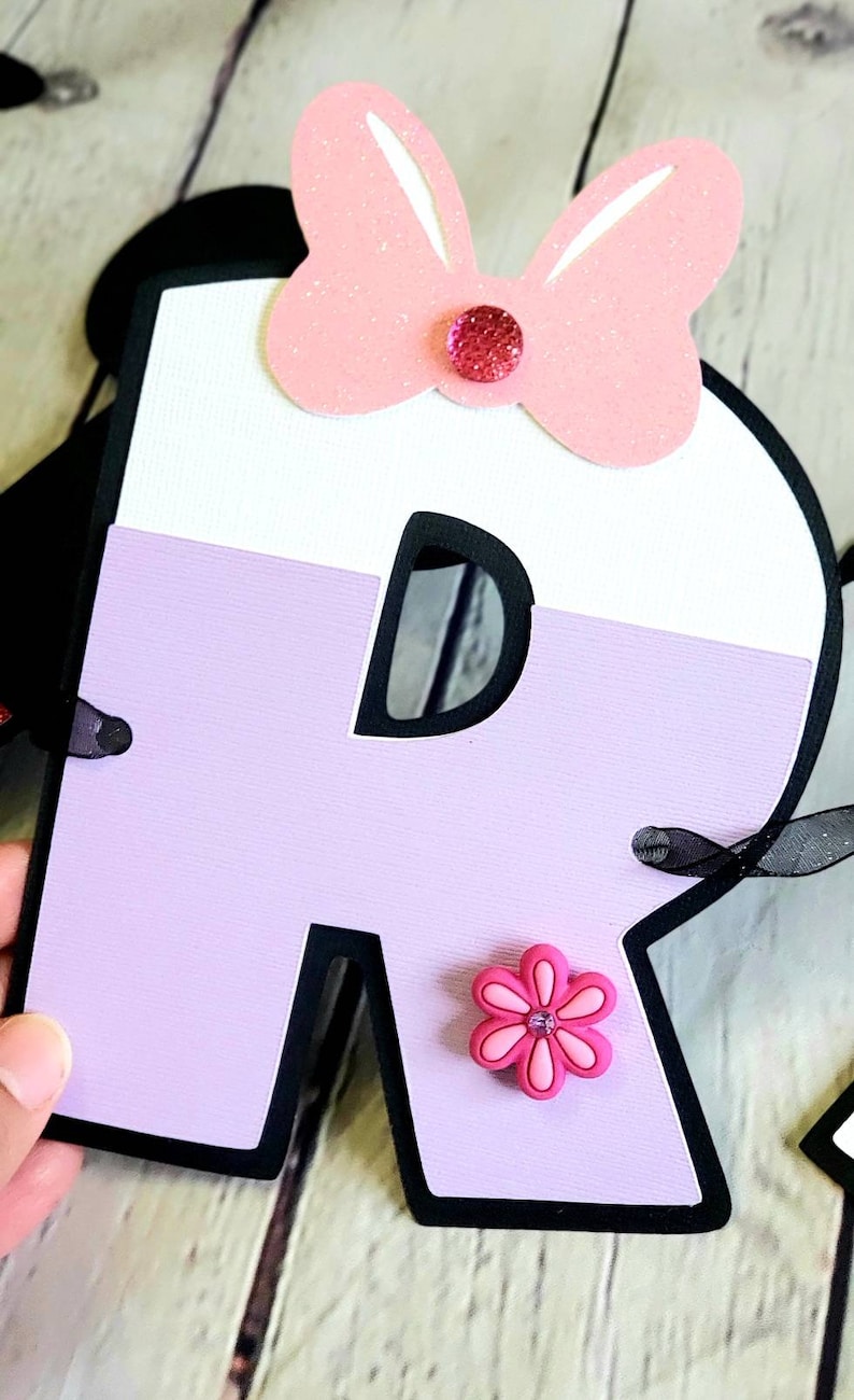 May include: A purple and white letter R with a black outline. The letter has a pink bow with a rhinestone in the center and a pink flower with a rhinestone in the center.