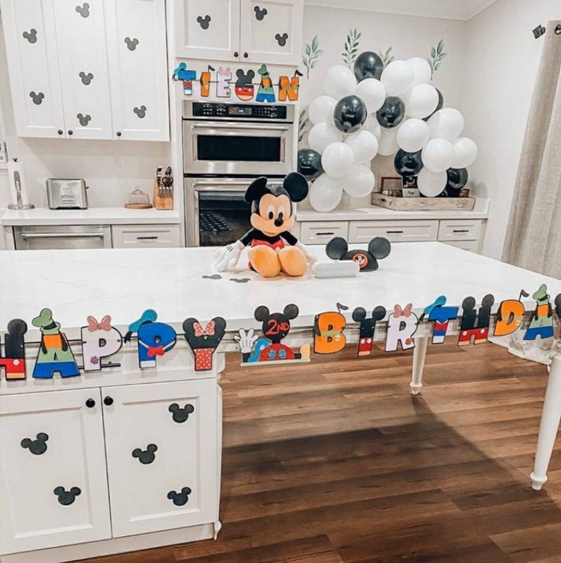 May include: A white kitchen island with a Mickey Mouse themed birthday banner that says "Happy Birthday". The banner is decorated with Mickey Mouse characters and the number "2". There is a Mickey Mouse plush toy sitting on the island.