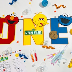 May include: A Sesame Street themed first birthday party banner with the letters O-N-E, featuring Elmo, Big Bird, and Cookie Monster. The banner is decorated with cookies, alphabet blocks, and a Sesame Street sign.