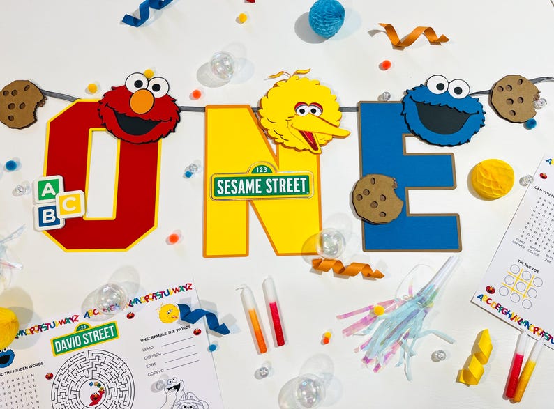 May include: A Sesame Street themed first birthday party banner with the letters O-N-E, featuring Elmo, Big Bird, and Cookie Monster. The banner is decorated with cookies, alphabet blocks, and a Sesame Street sign.