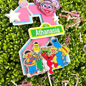 May include: A pink number two cake topper with a Sesame Street theme. The number two is decorated with a pink Big Bird character, a Sesame Street sign that says "Athanasia", and a group of Sesame Street characters including Elmo, Cookie Monster, Big Bird, Bert, Ernie, and Grover.