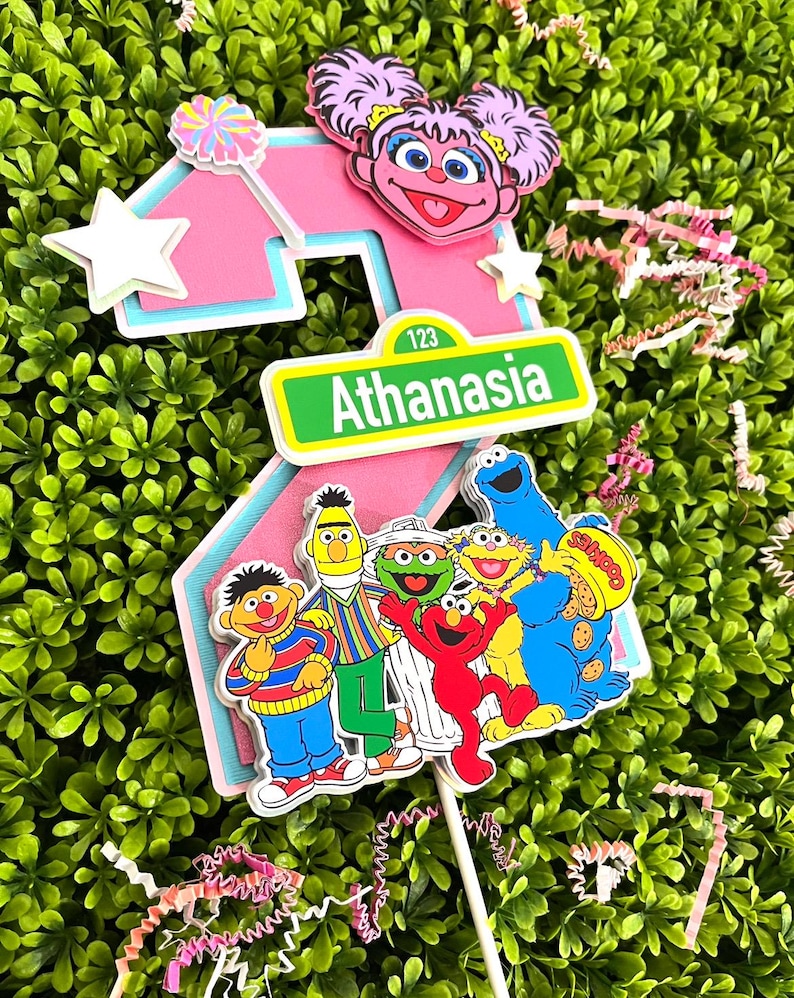 May include: A pink number two cake topper with a Sesame Street theme. The number two is decorated with a pink Big Bird character, a Sesame Street sign that says "Athanasia", and a group of Sesame Street characters including Elmo, Cookie Monster, Big Bird, Bert, Ernie, and Grover.