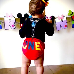 May include: A baby wearing a black shirt with suspenders and red and yellow "ONE"  shorts. The baby is standing in front of a colorful banner with the letters "A R N".