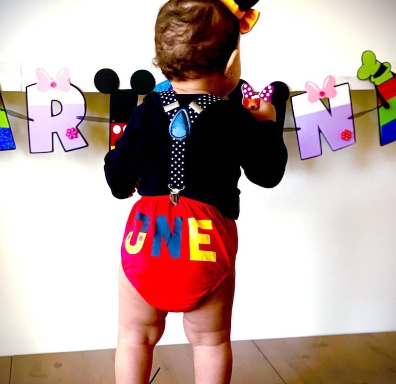 May include: A baby wearing a black shirt with suspenders and red and yellow "ONE"  shorts. The baby is standing in front of a colorful banner with the letters "A R N".