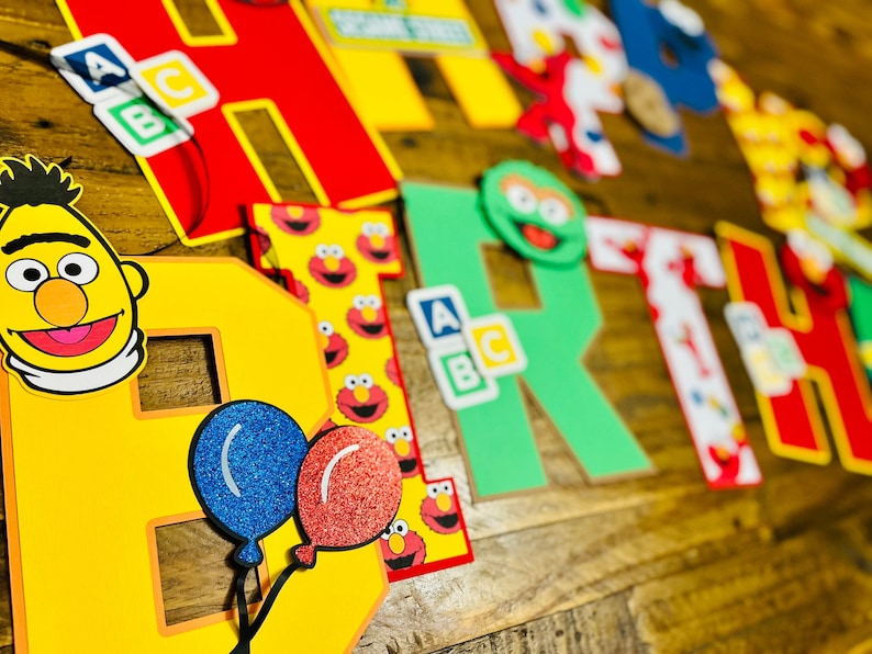 May include: A colorful "Happy Birthday" banner with Sesame Street characters, including Big Bird, Elmo, and Cookie Monster. The letters are large and made of cardboard. There are also small alphabet blocks with the letters A, B, and C.