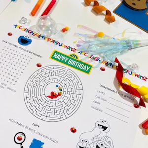 May include: A black and white printable activity sheet featuring Sesame Street characters Elmo, Cookie Monster, and Big Bird. The sheet includes a word search, a maze, a word scramble, and an I Spy game. The text on the sheet reads "Can you find the hidden words", "Happy Birthday", "Unscramble the words", "I Spy", and "How many Elmo's can you find?"