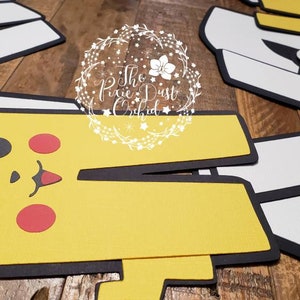 May include: A set of five yellow, red, white, and black felt cutouts of Pokemon characters. The characters are Pikachu, Charmander, Squirtle, Bulbasaur, and a trainer. The cutouts are designed to be used as party decorations or for crafts.