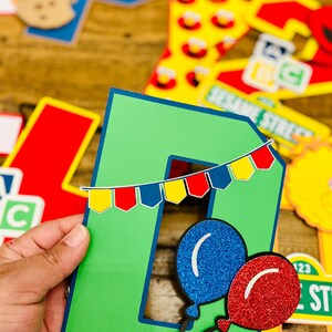 May include: Green letter D with a blue and red banner and two balloons, one blue and one red, with glitter. The letter is part of a Sesame Street themed banner.