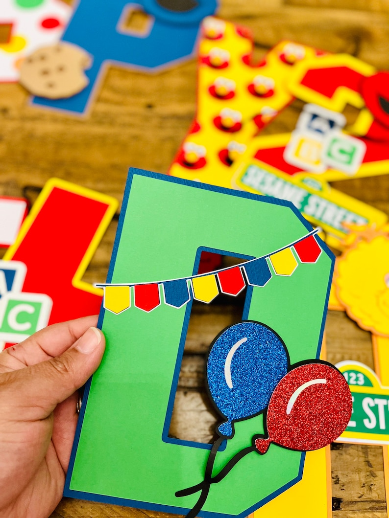 May include: Green letter D with a blue and red banner and two balloons, one blue and one red, with glitter. The letter is part of a Sesame Street themed banner.