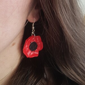 Könnte beinhalten: Rote Mohnblumen-Ohrringe mit schwarzem Mittelteil, hängend an silberfarbenen Haken. Die Ohrringe sind leuchtend rot und haben die Form einer Mohnblume. Das Thema der Ohrringe ist floral.