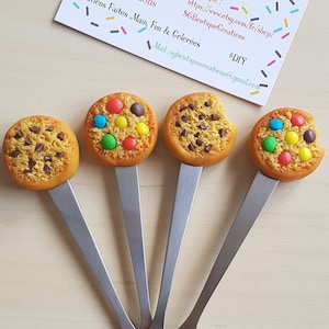 Peut inclure: Quatre cuillères en argent avec des dessus en forme de biscuits différents. Les biscuits sont décorés de pépites de chocolat, de vermicelles et de bonbons colorés. Les cuillères sont disposées en éventail sur une surface en bois clair.