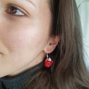 Puede incluir: Pendientes de taza roja con lunares blancos y una cucharada de crema batida blanca en la parte superior. Los pendientes cuelgan de la oreja de una persona.