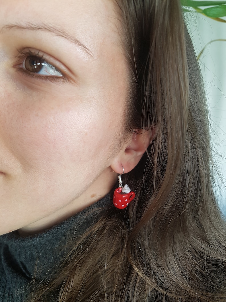 Puede incluir: Pendientes de taza roja con lunares blancos y una cucharada de crema batida blanca en la parte superior. Los pendientes cuelgan de la oreja de una persona.