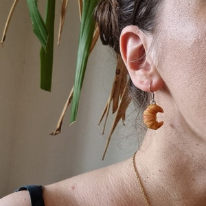 Puede incluir: Un par de pendientes de oro con un dije de croissant. Los pendientes son usados por una persona con cabello casta&ntilde;o.