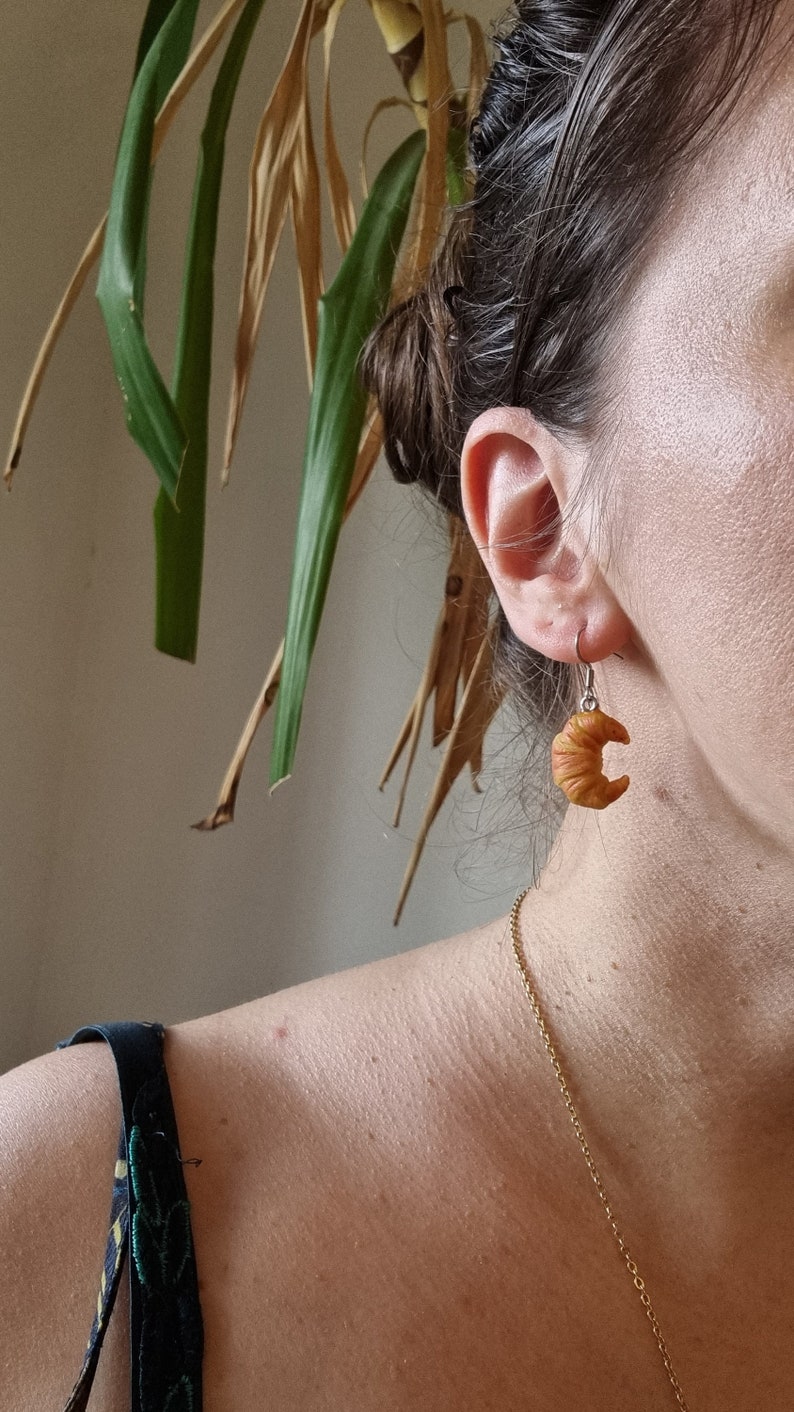 Puede incluir: Un par de pendientes de oro con un dije de croissant. Los pendientes son usados por una persona con cabello casta&ntilde;o.