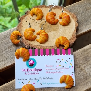 Puede incluir: Pendientes en forma de croissant hechos de arcilla polim&eacute;rica. Los pendientes son de color marr&oacute;n claro con un tono amarillo. Los pendientes cuelgan de ganchos plateados. Los pendientes se muestran sobre una superficie de madera con una tarjeta de visita que dice "SGBoutique & Creations" y "Creations Faites Main, Fun & Celer&eacute;es".