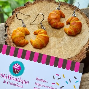Puede incluir: Un par de pendientes en forma de croissant hechos de arcilla polim&eacute;rica. Los pendientes son de color marr&oacute;n claro con un tono dorado. Los pendientes cuelgan de ganchos plateados. Los pendientes est&aacute;n sobre una superficie de madera. La imagen tambi&eacute;n incluye una tarjeta de visita con el texto "SGBoutique & Cr&eacute;ations" y una direcci&oacute;n de sitio web.