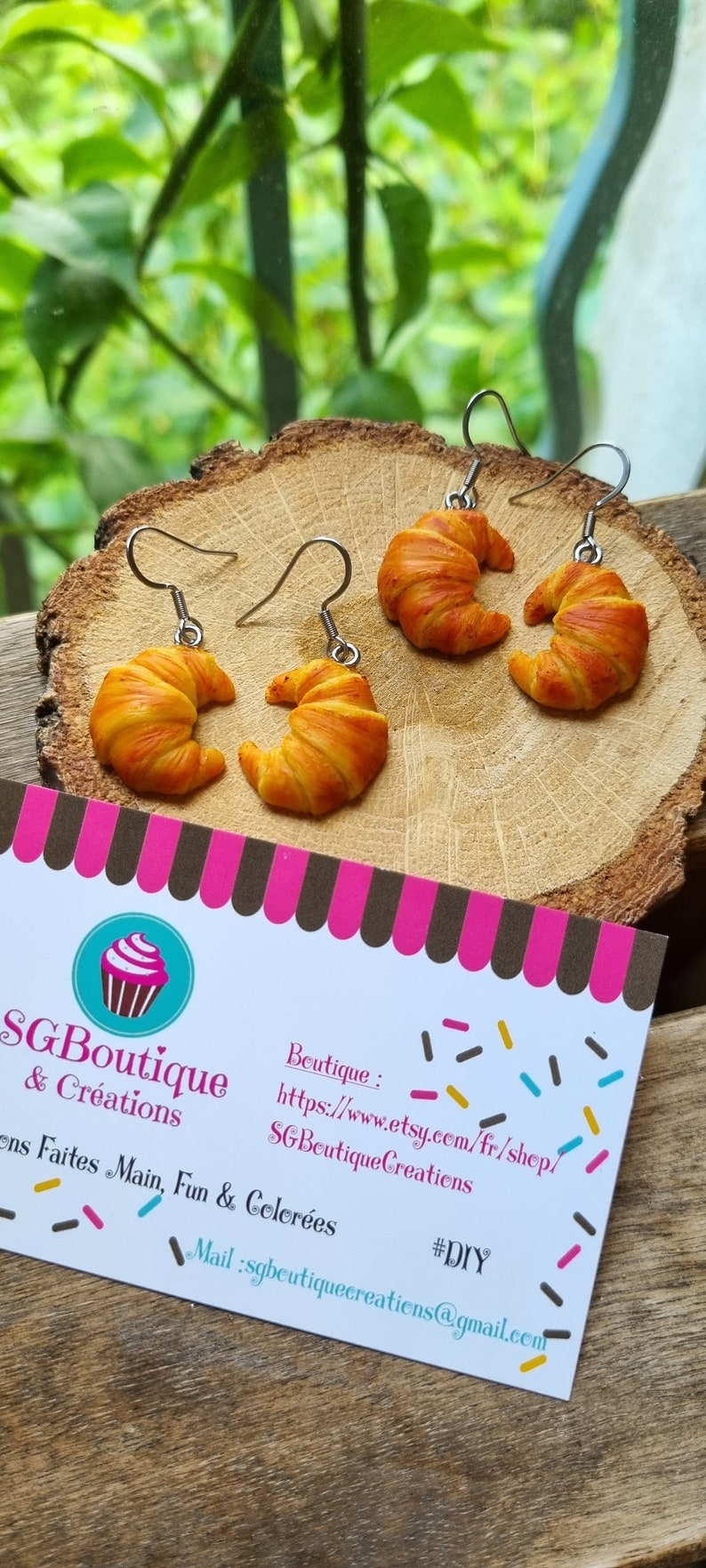 Puede incluir: Un par de pendientes en forma de croissant hechos de arcilla polim&eacute;rica. Los pendientes son de color marr&oacute;n claro con un tono dorado. Los pendientes cuelgan de ganchos plateados. Los pendientes est&aacute;n sobre una superficie de madera. La imagen tambi&eacute;n incluye una tarjeta de visita con el texto "SGBoutique & Cr&eacute;ations" y una direcci&oacute;n de sitio web.