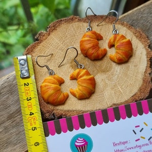 Puede incluir: Un par de pendientes con forma de croissant con una regla amarilla para referencia de tama&ntilde;o. Los pendientes est&aacute;n hechos de arcilla polim&eacute;rica y miden aproximadamente 5 cm de largo. La regla es amarilla con marcas negras. Los pendientes est&aacute;n sobre una superficie de madera con una tarjeta de visita que dice "SG Boutique & Creations" y "Cr&eacute;ations Faites Main, Fun & Celer&eacute;es".