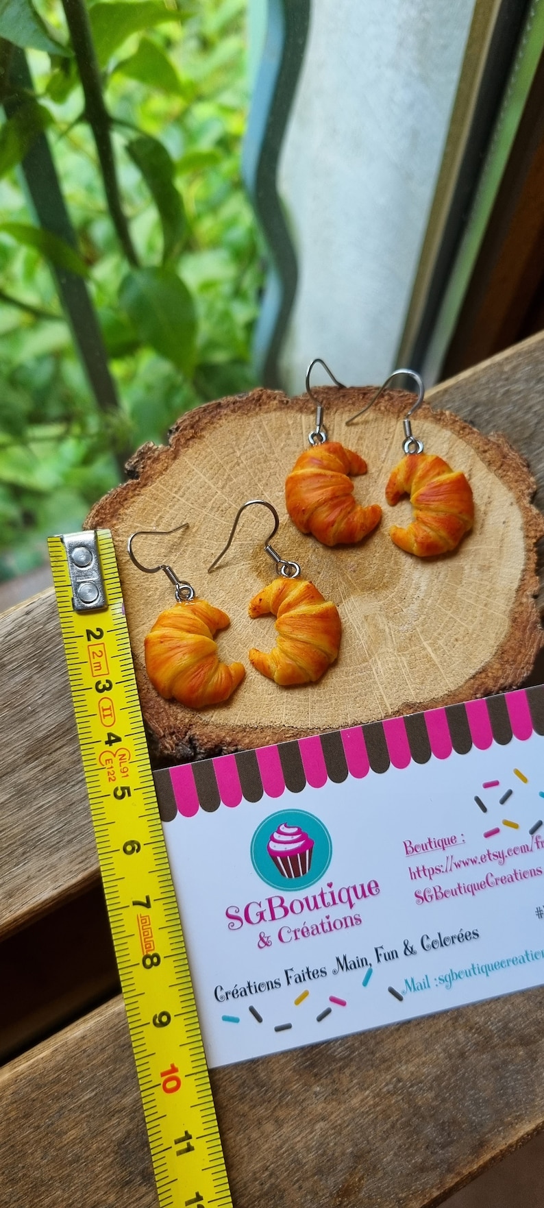 Puede incluir: Un par de pendientes con forma de croissant con una regla amarilla para referencia de tama&ntilde;o. Los pendientes est&aacute;n hechos de arcilla polim&eacute;rica y miden aproximadamente 5 cm de largo. La regla es amarilla con marcas negras. Los pendientes est&aacute;n sobre una superficie de madera con una tarjeta de visita que dice "SG Boutique & Creations" y "Cr&eacute;ations Faites Main, Fun & Celer&eacute;es".