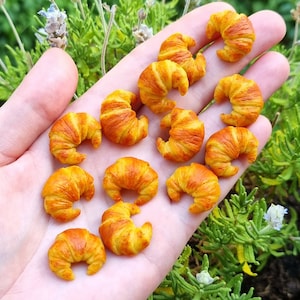 Puede incluir: Una mano sosteniendo una colecci&oacute;n de mini croissants hechos de arcilla polim&eacute;rica. Los croissants son de color amarillo claro con acentos marrones.