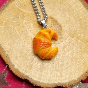 Puede incluir: Un collar de cadena plateada con un colgante de croissant. El croissant est&aacute; hecho de arcilla polim&eacute;rica amarilla y naranja y tiene forma de media luna.
