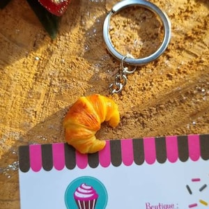 Puede incluir: Un llavero con un peque&ntilde;o charm con forma de croissant de aspecto realista. El charm es de color marr&oacute;n claro con un contorno marr&oacute;n ligeramente m&aacute;s oscuro. El llavero est&aacute; unido a un anillo y una cadena de metal plateado. El llavero se muestra sobre una superficie de madera con un fondo a rayas rosa y negro.