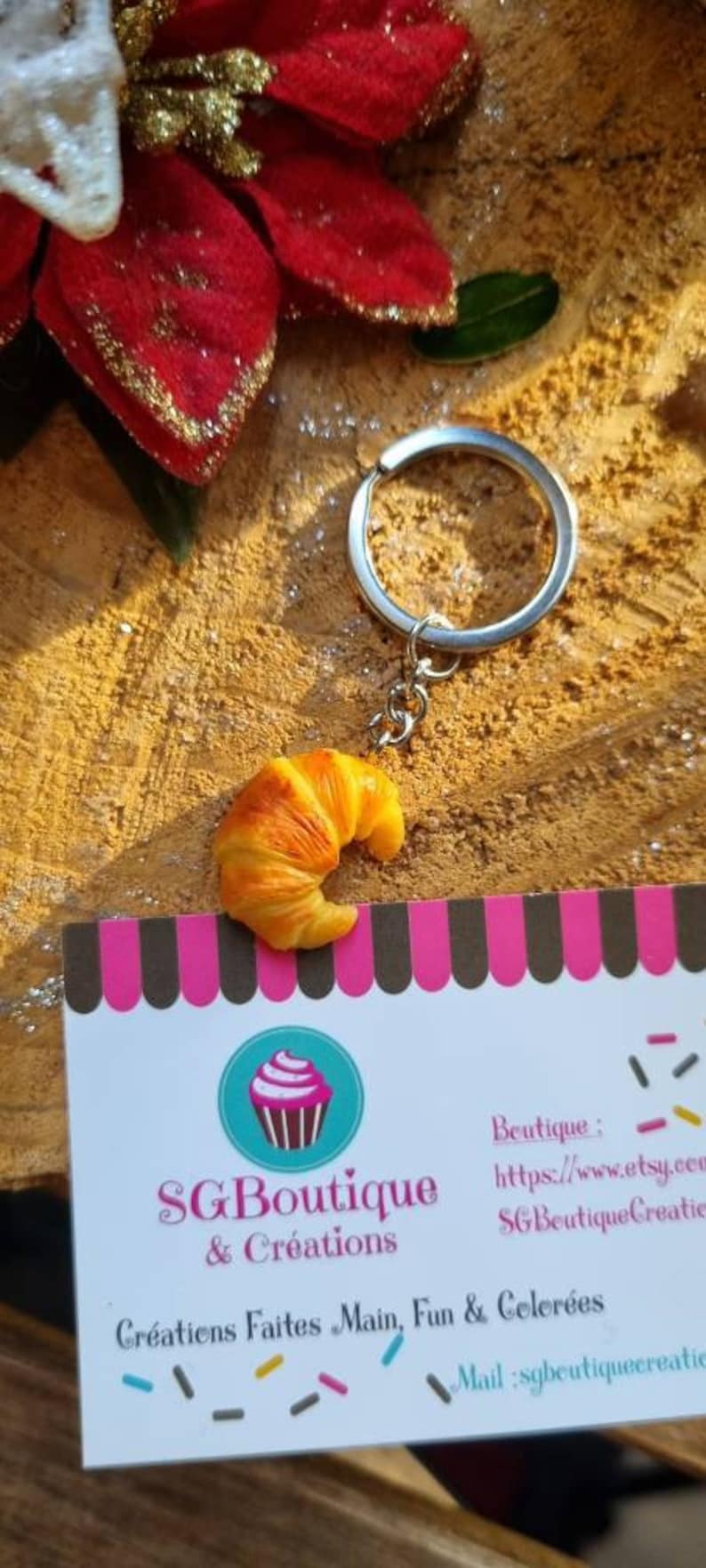 Puede incluir: Un llavero con un peque&ntilde;o charm con forma de croissant de aspecto realista. El charm es de color marr&oacute;n claro con un contorno marr&oacute;n ligeramente m&aacute;s oscuro. El llavero est&aacute; unido a un anillo y una cadena de metal plateado. El llavero se muestra sobre una superficie de madera con un fondo a rayas rosa y negro.